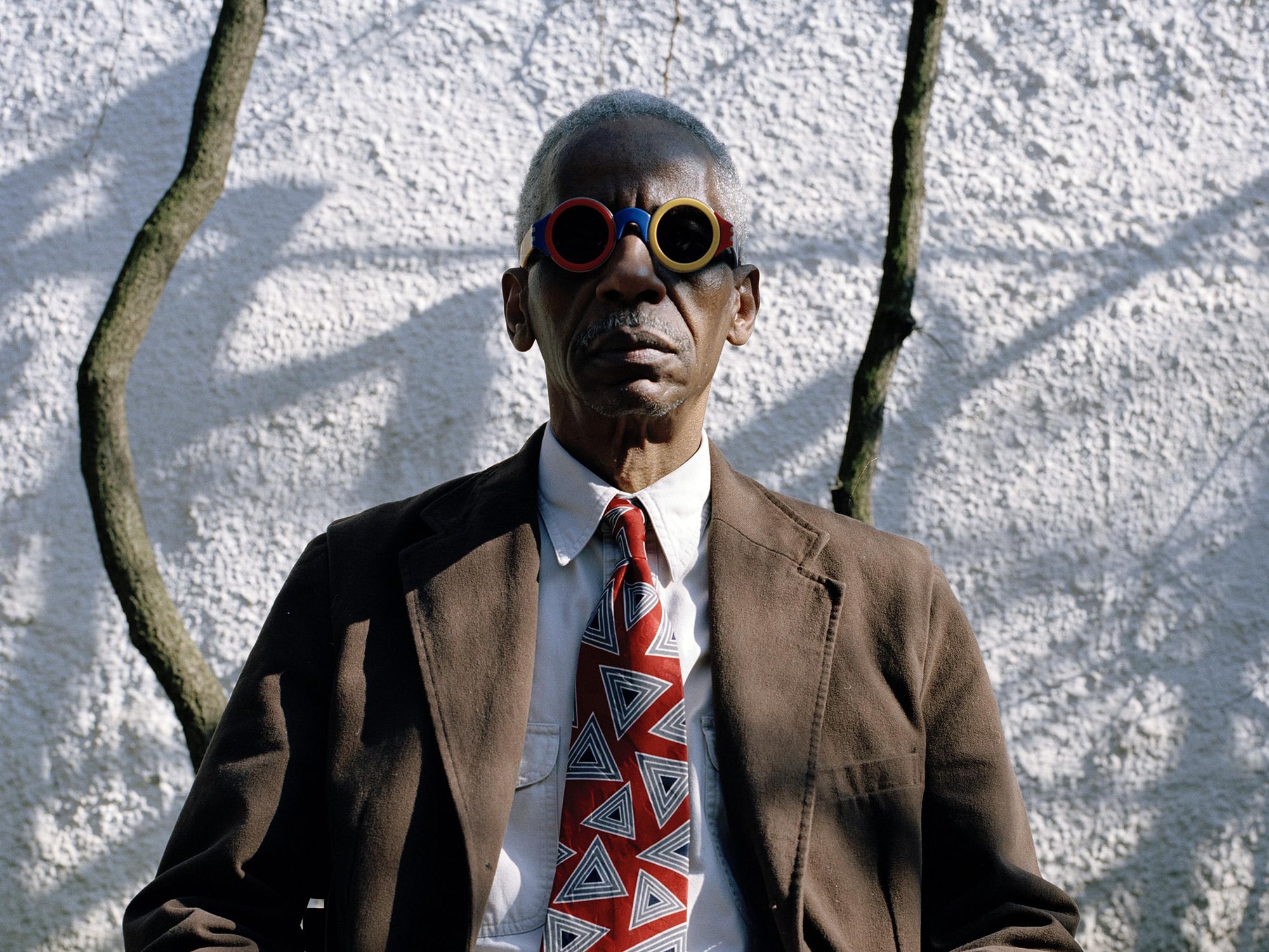A photo shows musician Roscoe Mitchell seated in front of a stucco wall with a tree between him and the wall. He is wearing goggles with dark lenses and colorful frames, a brown jacket, a red tie, and a white dress shirt.