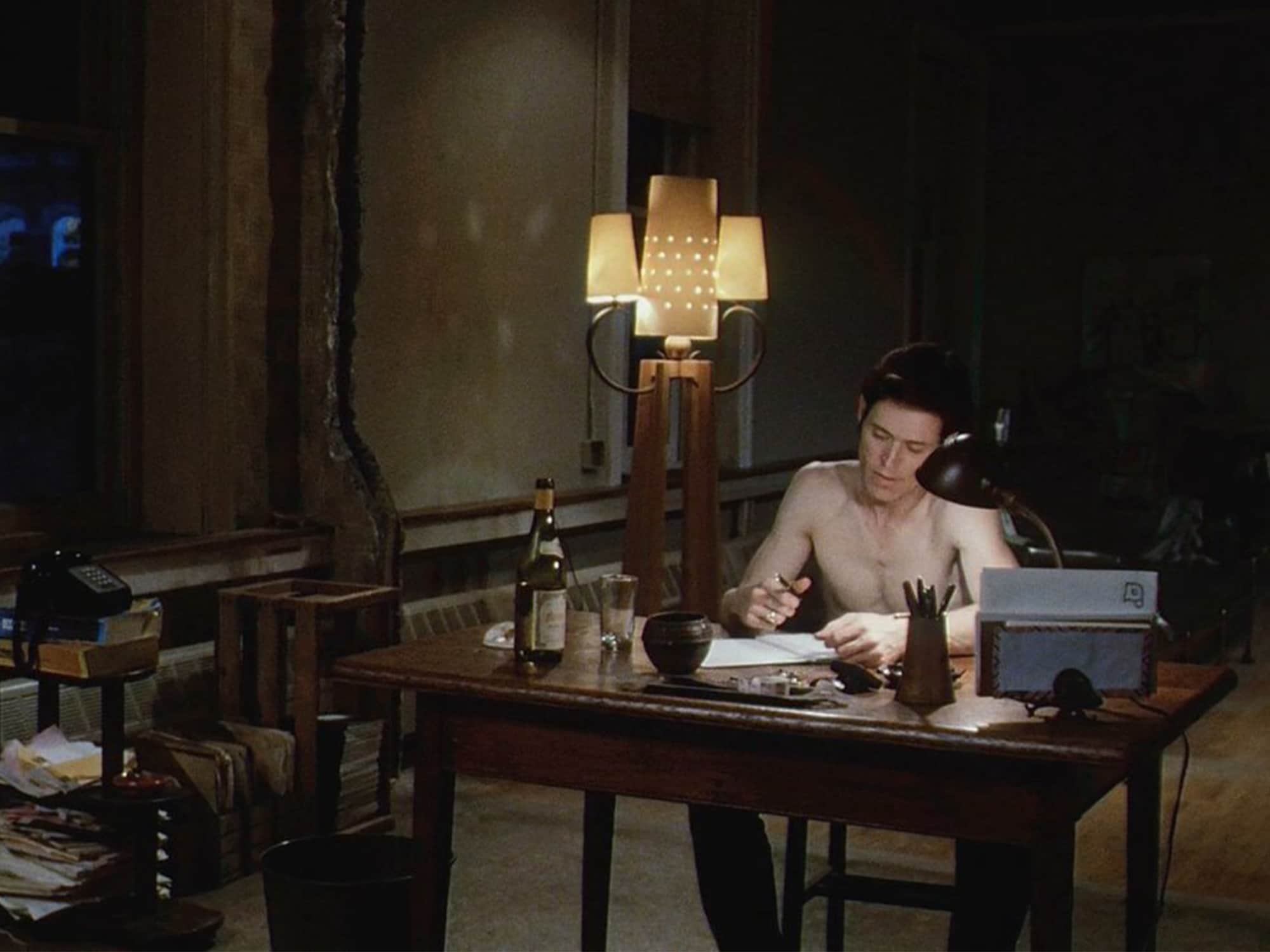 John LeTour (Willem Dafoe) is shown writing at a desk in his largely empty apartment at night, shirtless. There is a tall, stylized lamp behind him and a wine bottle sitting on the table next to a glass.