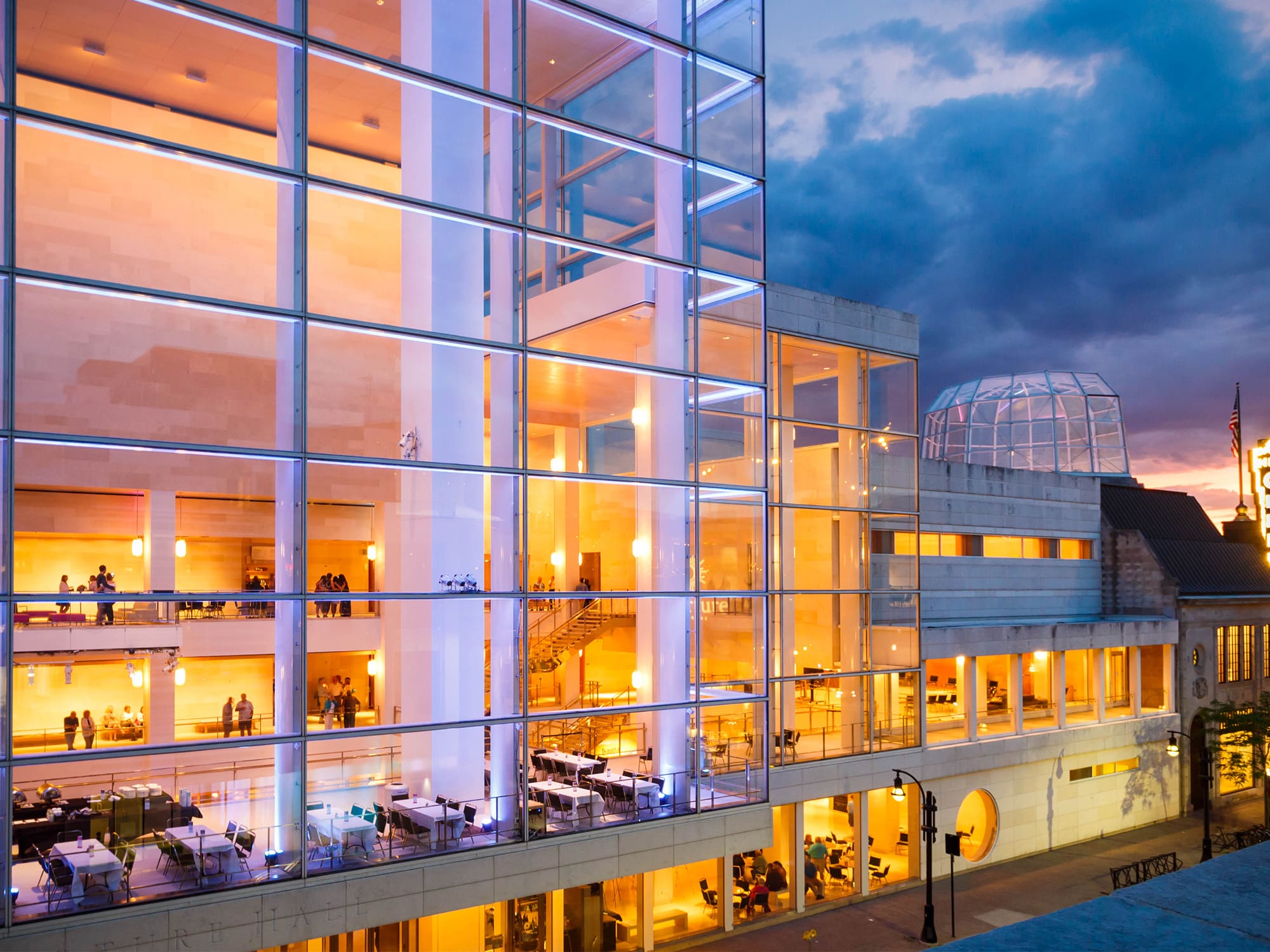 A photo shows the side of the Overture Center that faces out onto North Fairchild Street in downtown Madison. The multi-story, glass facade reveals people gathering in a lobby outside of Overture Hall, and warm lighting inside. In the background is an evening sky and, because of the angle of the photo, a view of the glass dome that tops Overture's rotunda.