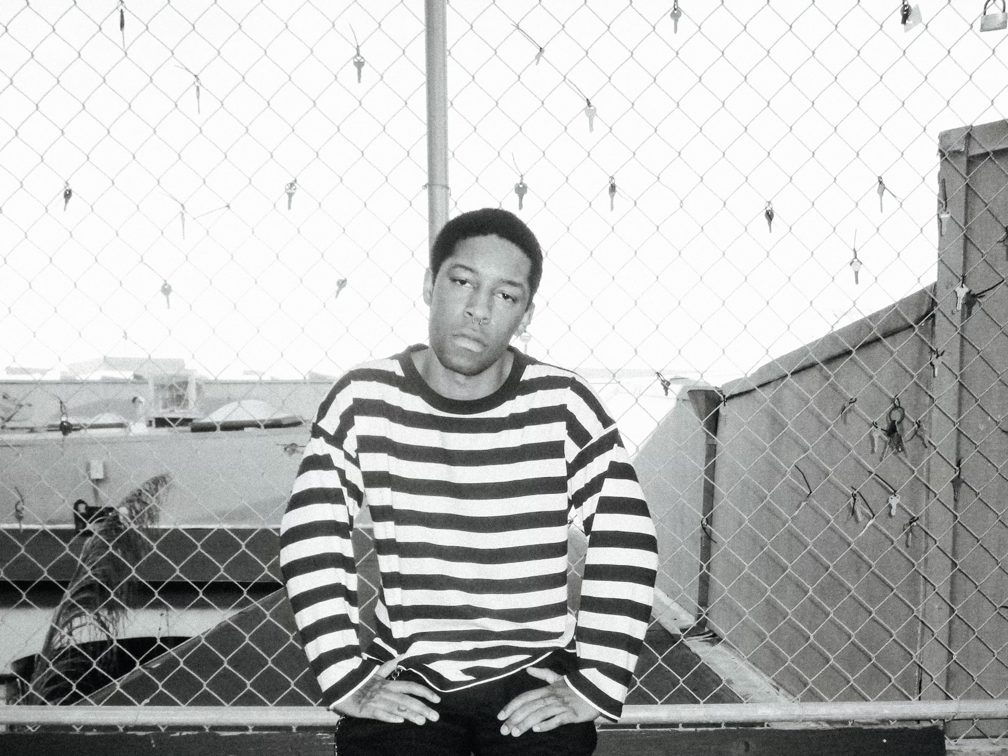 A black-and-white photo shows musician KennyHoopla sitting in front of a chain-link fence.