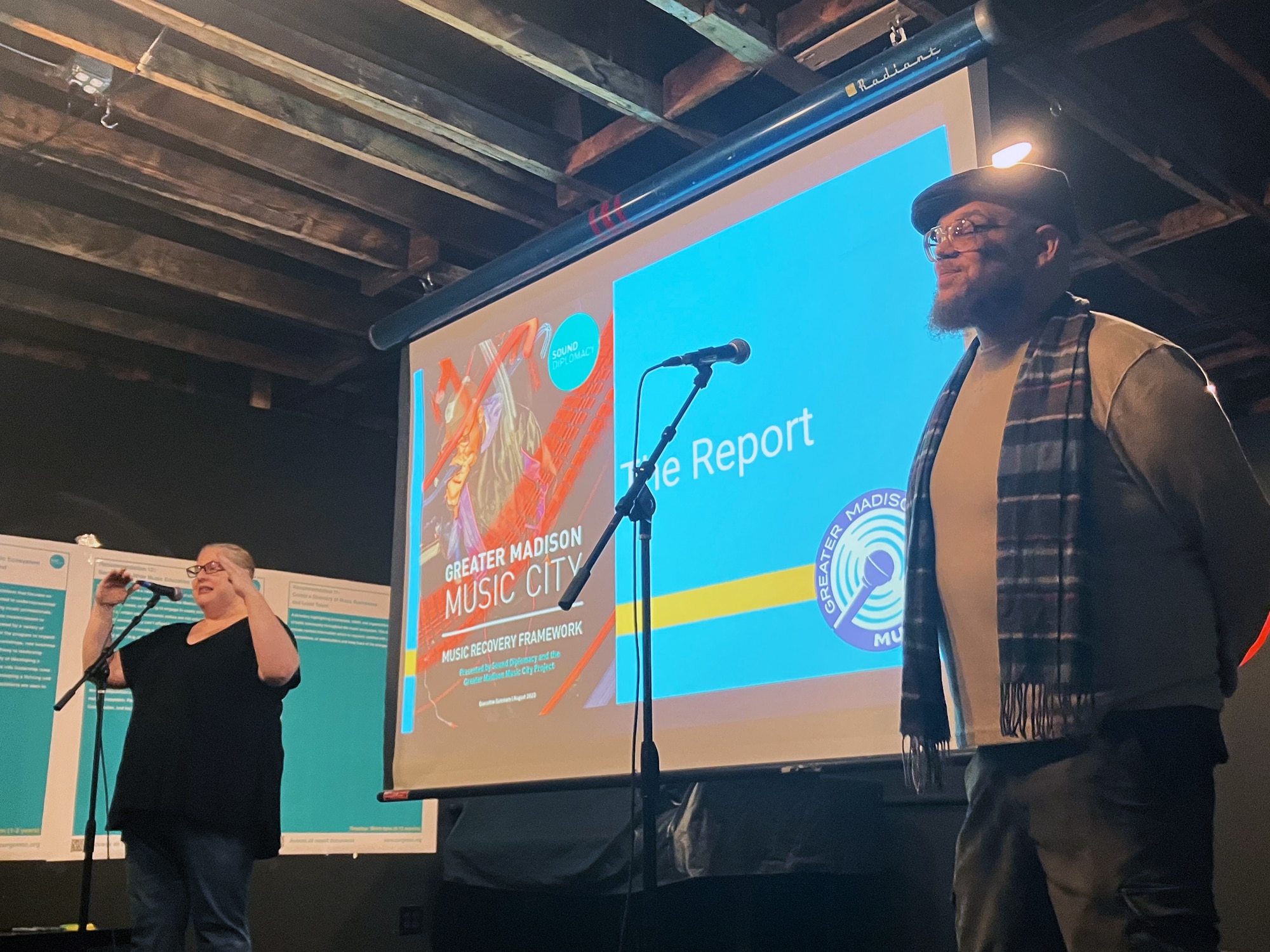 A photo shows Karen Reece, left, and Rob Franklin standing on the slightly elevated stage at Café Coda, speaking into microphones. Behind them is a projection screen showing the cover image of the report.