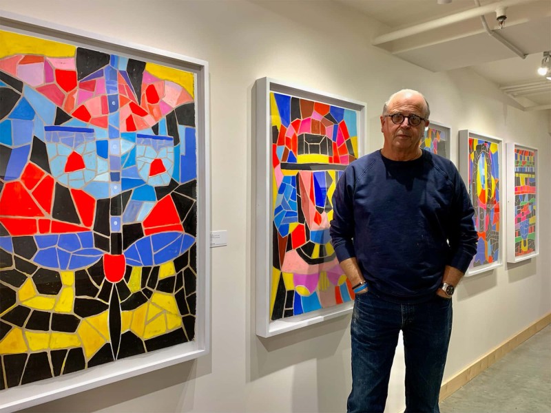 A photo shows artist T.L. Solien standing in front of his works in a gallery. The works are mosaic-like paintings comprising multicolored fragments that suggest shapes and patterns.