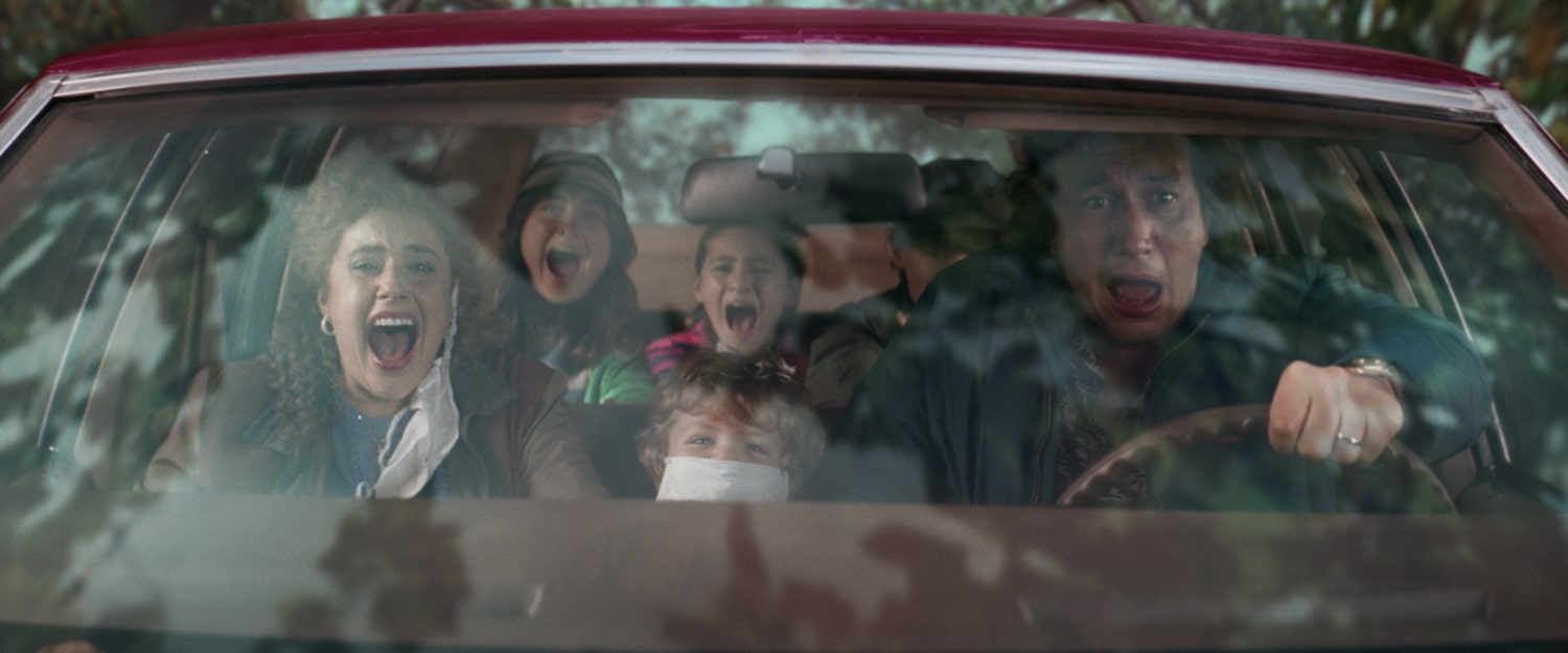 Babette Gladney (Greta Gerwig), Jack Gladney (Adam Driver), and their three children scream while in their red station wagon. The youngest Wilder (Henry Moore) sits masked between Jack and Babette in the front seat looking amused.