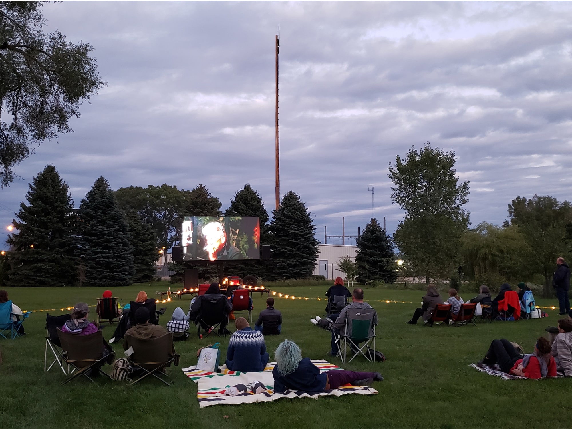 A photo shows a screening of "David Lynch: The Art Life" on a lawn outside of Tandem Press in September 2022. The audience is spread across the grass, sitting on lawn chairs and blankets. They are shown from behind, facing the screen, on which a shot of David Lynch smoking a cigarette is visible. 