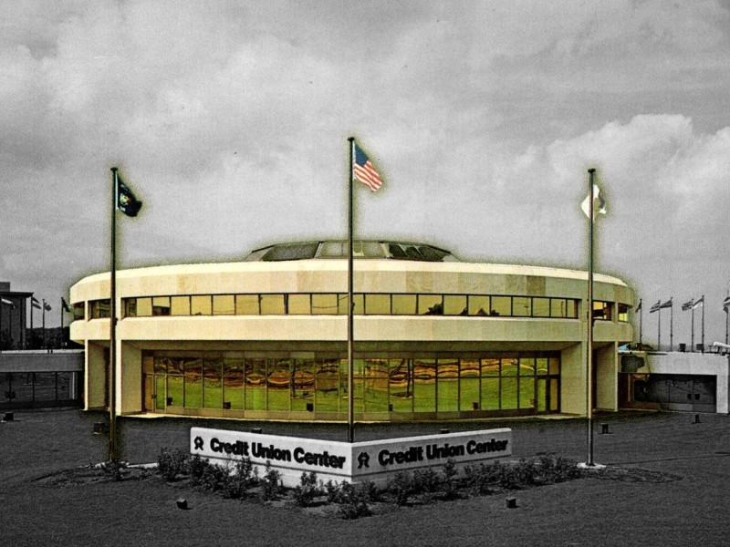 A photo illustration shows the now-demolished circular headquarters of CUNA Mutual Group on Mineral Point Road. The background of the office campus is set in black and white while the building is foregrounded in color.