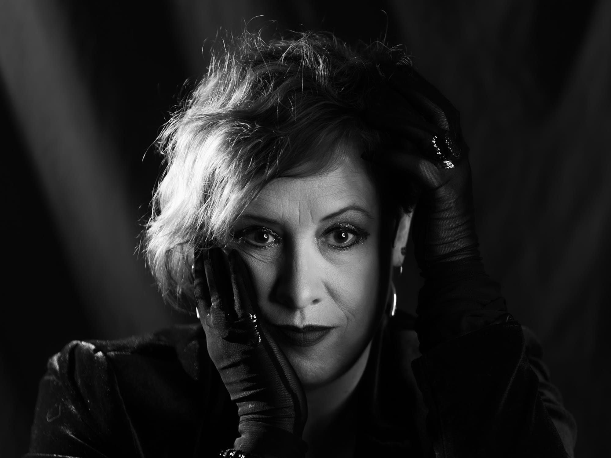 A black-and-white studio photo shows Jen Wilson facing the camera, wearing black gloves with her face resting on one hand and her other hand reaching up toward her hair.