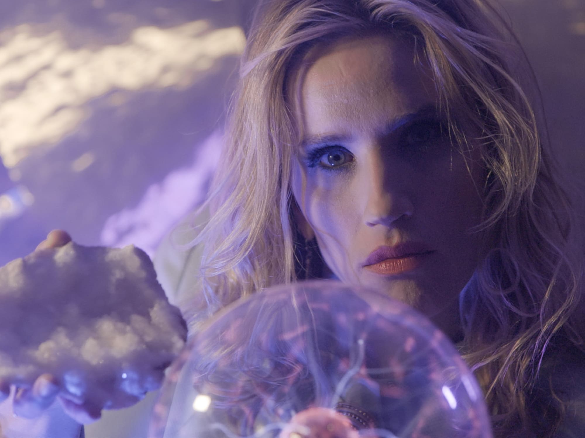 A still from Cribshitter's "Crystal City" video shows Christine Christenson holding a large crystal in one hand while gazing into a plasma ball toy positioned between her face and the camera. The scene is tinted with purple light, and behind Christenson is a wall with a large cymbal or gong mounted on it. 