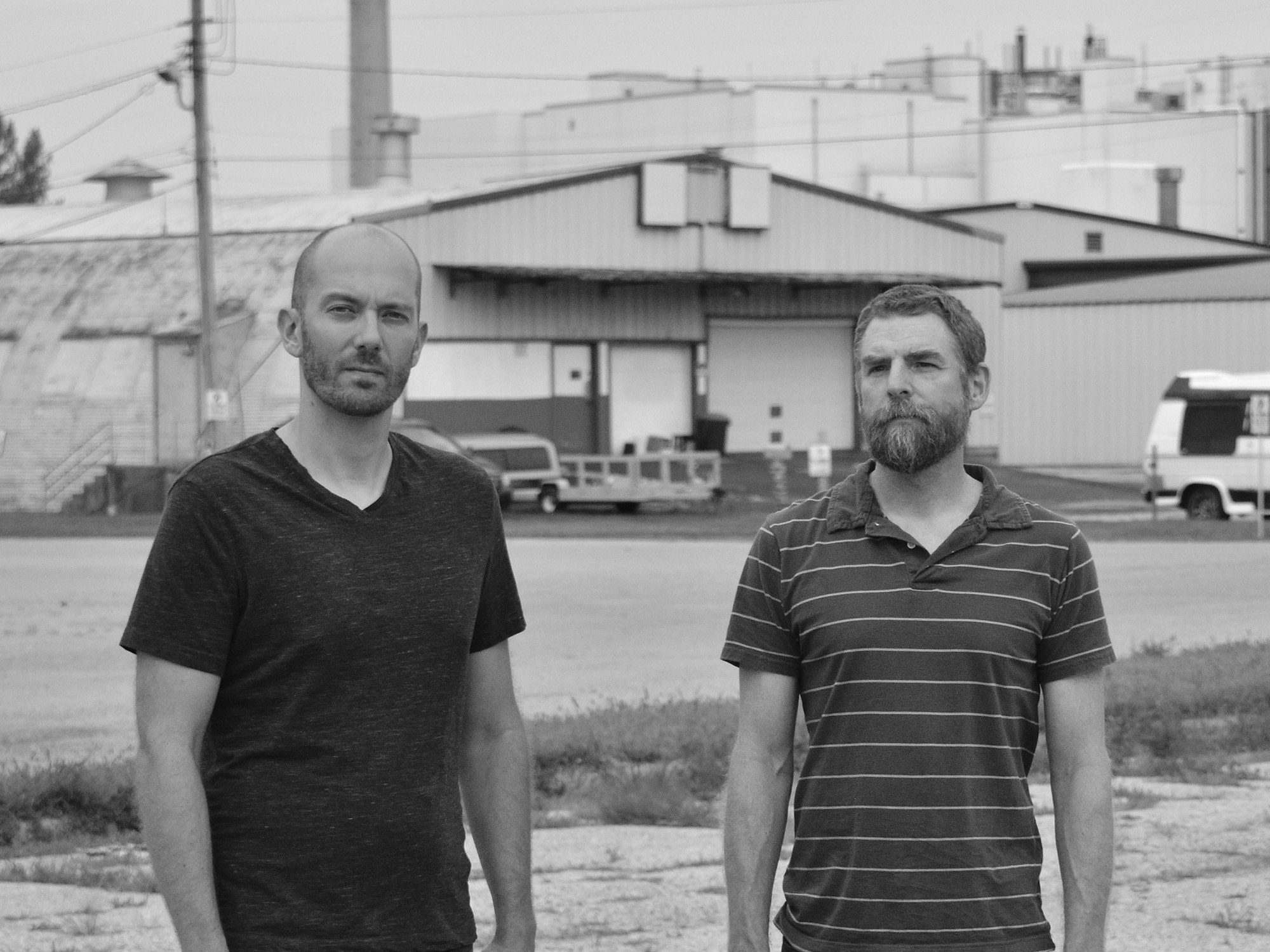 A black-and-white photo shows musicians Tony Barba (left) and Michael Brenneis standing next to each other and facing the camera. In the background is a nondescript industrial building with a loading dock.