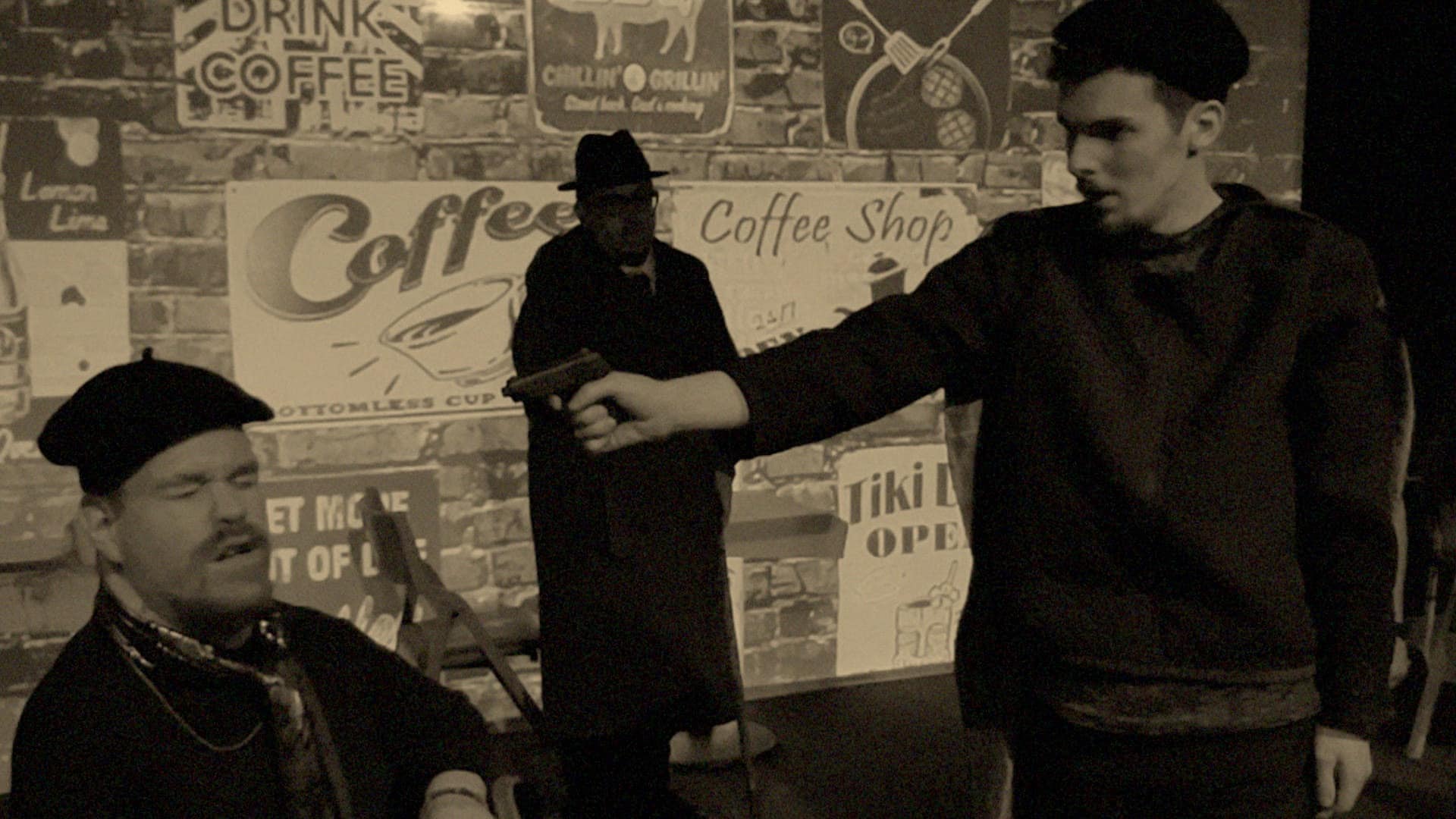 A scene from Heather Renken's "Elephant In The Room" that features three actors. Rikki Christman (right) points a toy gun at Joseph Wahlers (left, seated). James Burreson stands cloaked in the shadows near the center of the frame in front of a vintage coffee shop backdrop.
