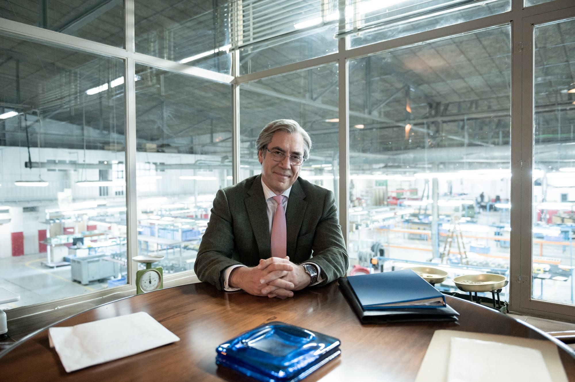 Blanco (Javier Bardem) smiles as he sits at a table in his office that overlooks the factory floor of Blanco Scales.