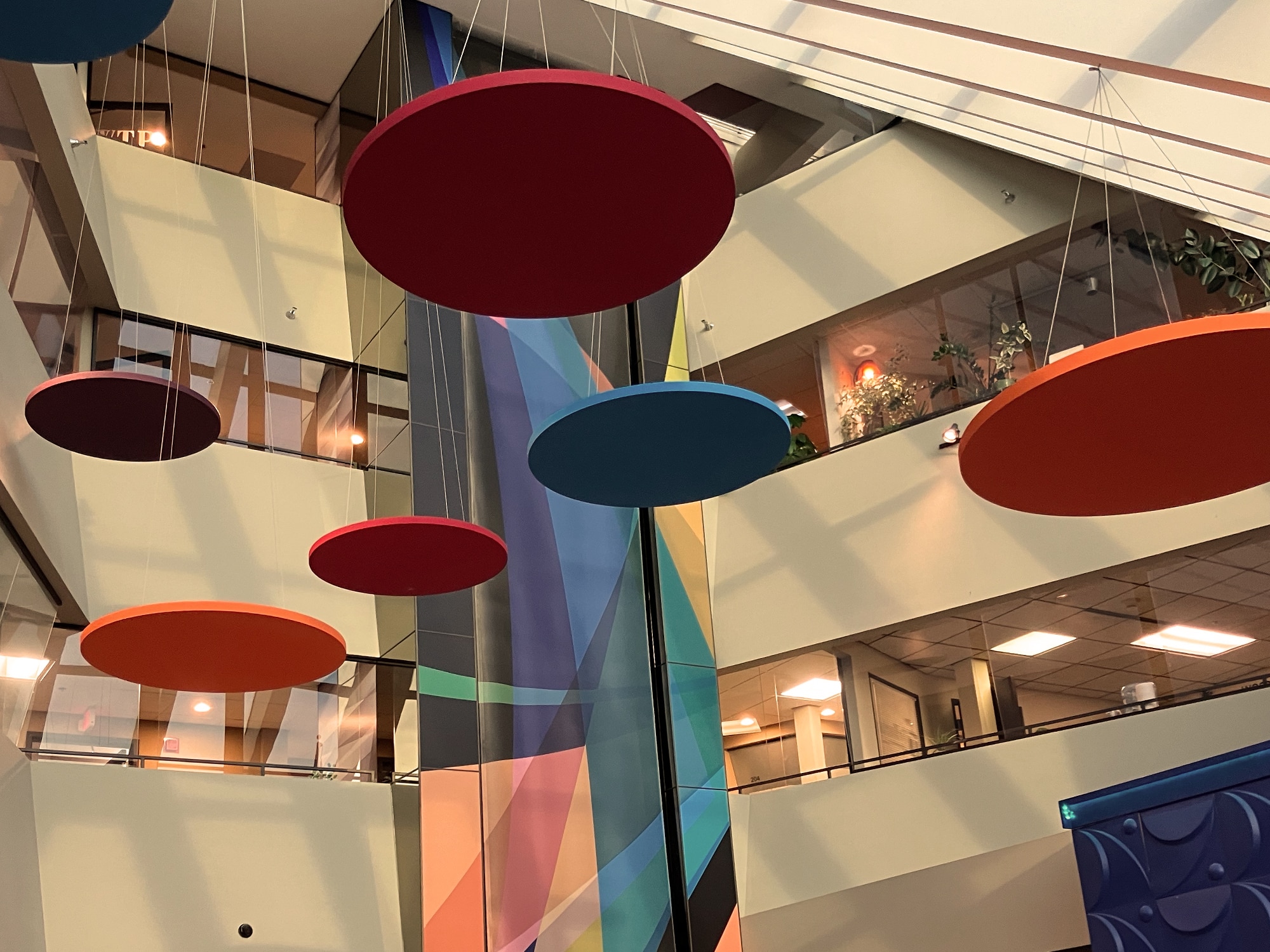 The Water Tower Place lobby atrium, seen from floor level, facing inward toward the offices in the buildings. Multiple large, brightly colored discs hang suspended from the ceiling, and a long, multicolored fabric hanging spans an elevator shaft that runs down the middle of several levels of office space.