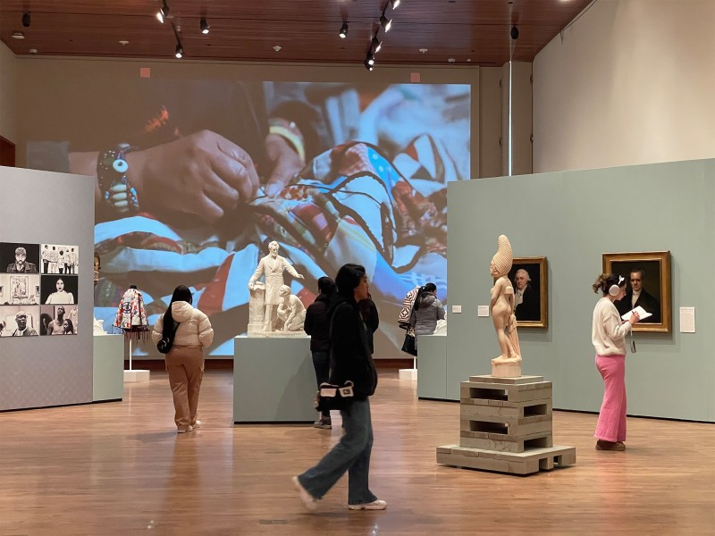 A view of the "re:mancipation" exhibition shows several visitors walking amongst paintings, sculptures and other pieces in a gallery at the Chazen Museum of Art. Behind them is a large wall on which an image of a pair of hands weaving a quilt is projected.