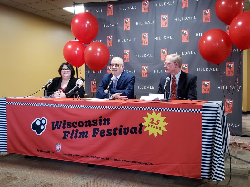 Hilldale General Manager Nanci Horn (left), Wisconsin Film Festival Director Of Operations Ben Reiser (center), and District 11 Alder Bill Tishler (right) speak at a Tuesday morning press conference in the lobby of the former AMC Madison 6. A "Wisconsin Film Festival 25 years" banner adorns the table with red balloons and "WFF Hilldale" logo backdrop.