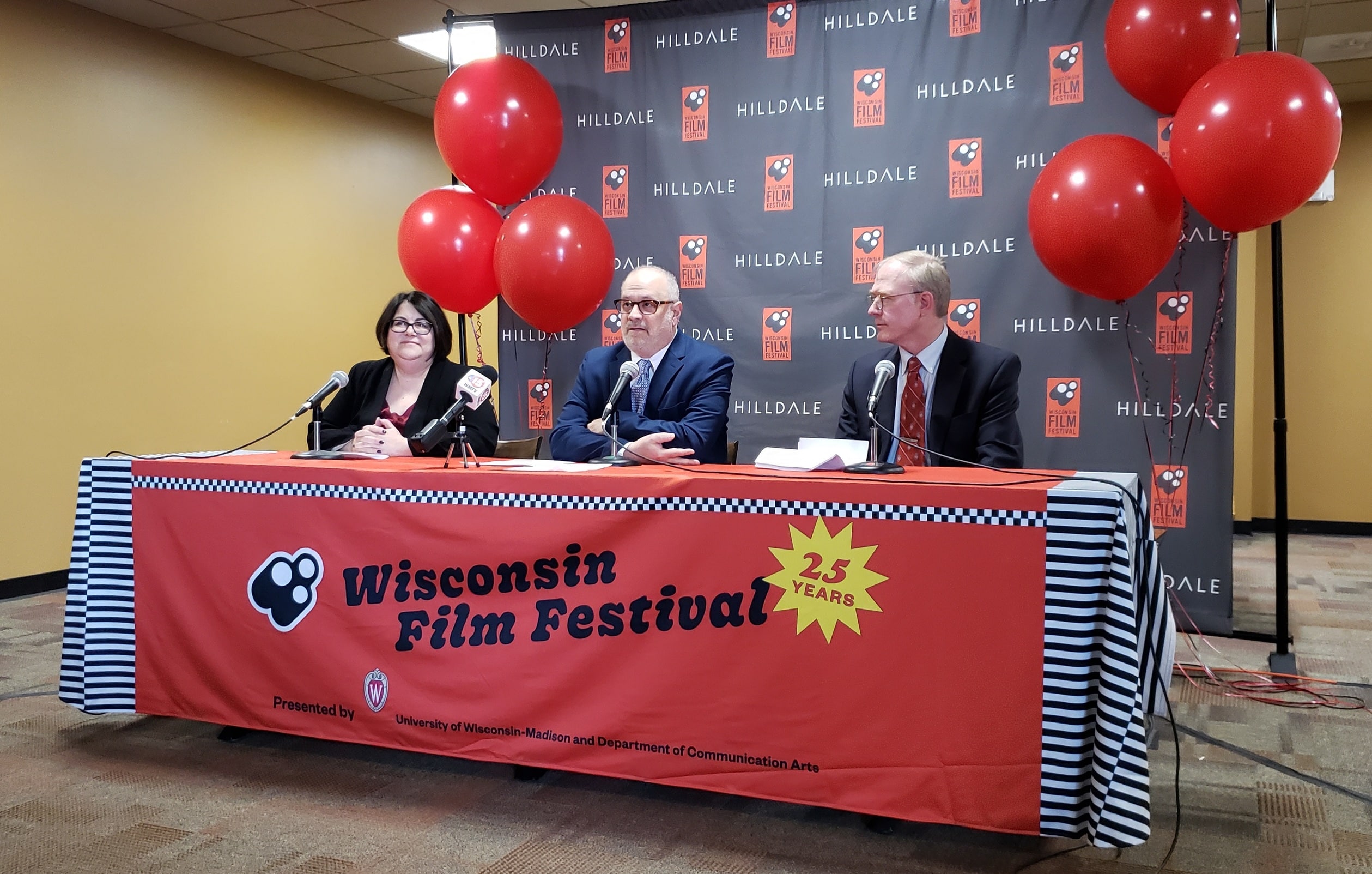 Hilldale General Manager Nanci Horn (left), Wisconsin Film Festival Director Of Operations Ben Reiser (center), and District 11 Alder Bill Tishler (right) speak at a Tuesday morning press conference at the former AMC Madison 6. A "Wisconsin Film Festival 25 years" banner adorns the table with red balloons and "WFF Hilldale" logo backdrop.