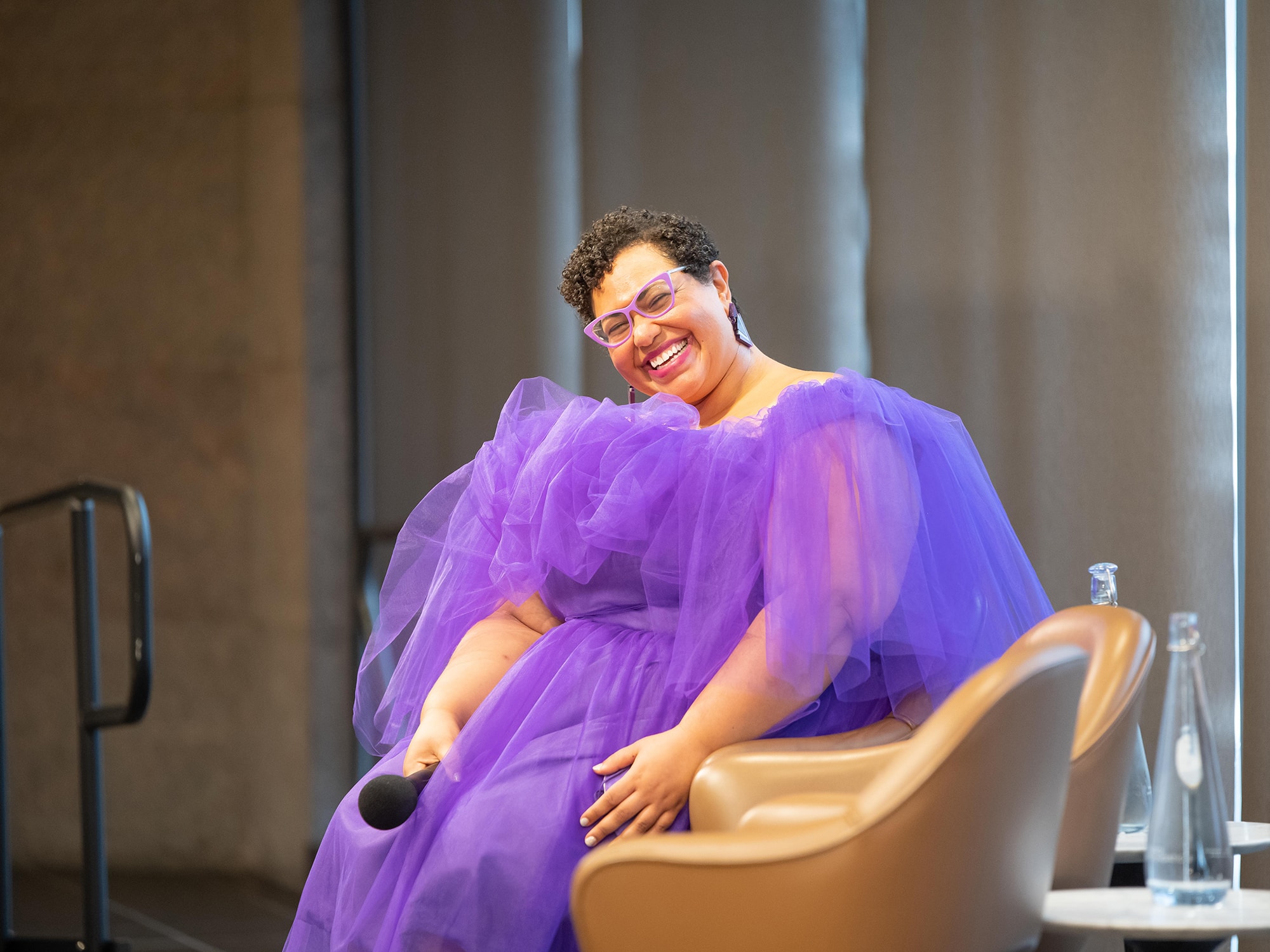 Dr. Sami Schalk is shown seated in a chair on a stage during a recent book tour. She is holding a microphone in one hand, and is smiling towards the camera as she wears a fluffy purple dress.