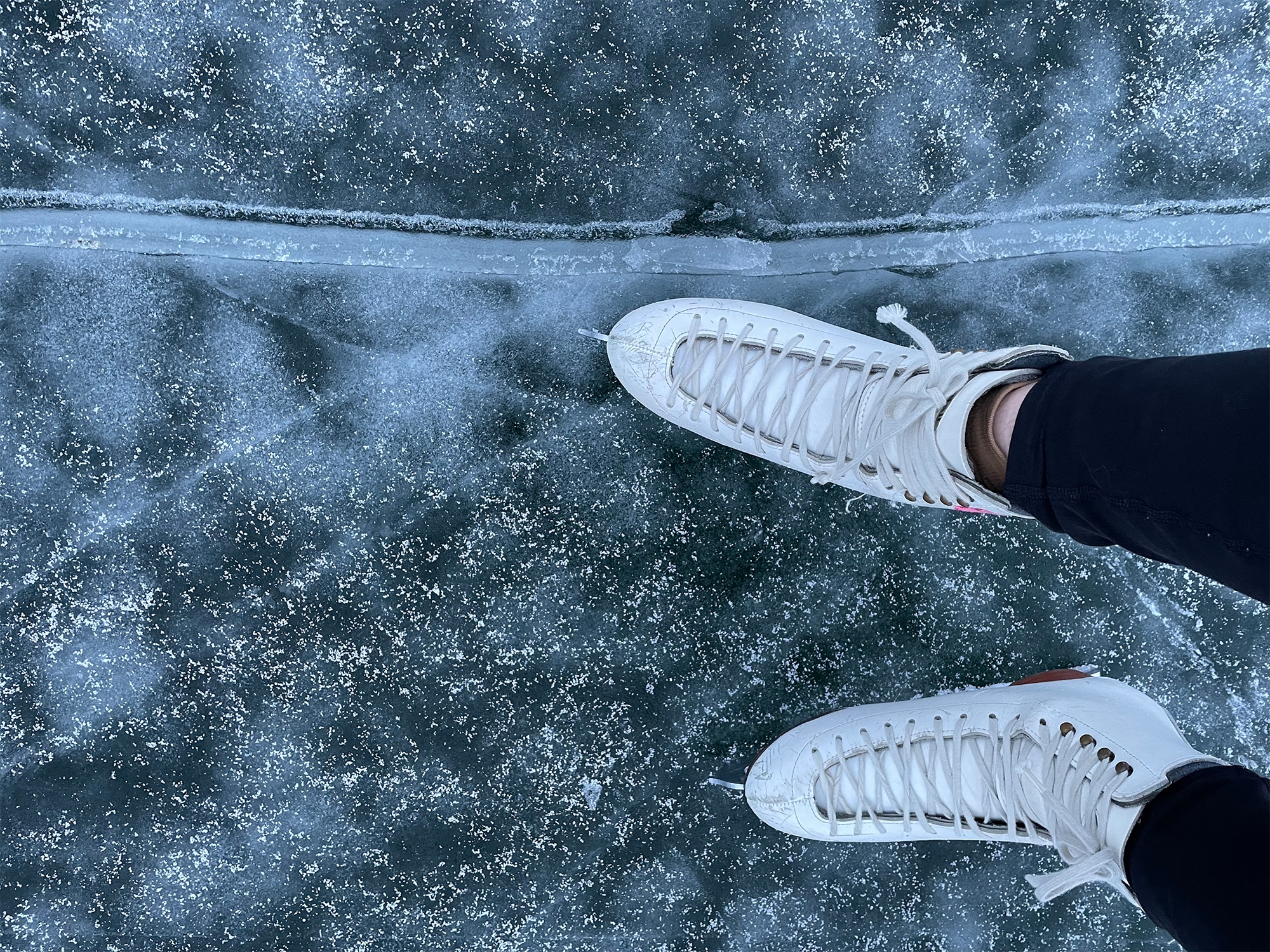 A photo taken from the perspective of an ice skater looking downward shoes a pair of skates and the frozen lake ice below it.