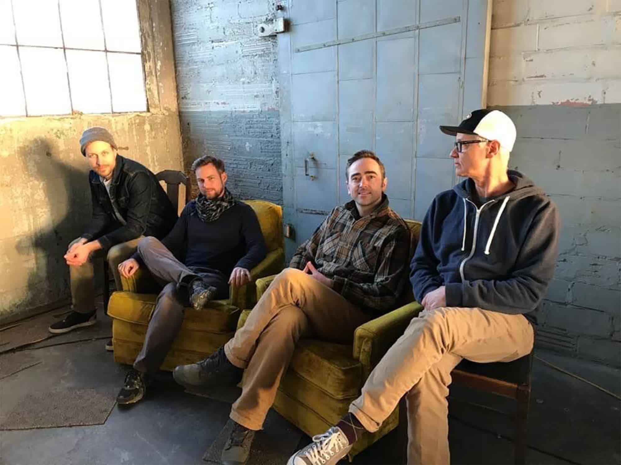 The four members of Disaster Passport are shown sitting on chairs in an industrial building, with a brick wall and large sliding door behind them.