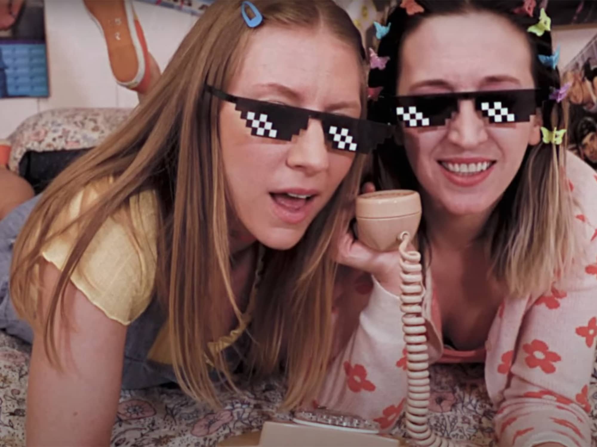 A still of Seasaw's "Dial Me Up" video in which the members are huddled together on a bed, smiling and holding up a landline telephone. Each has a pair of sunglasses superimposed onto their face by way of a lo-fi video effect. 