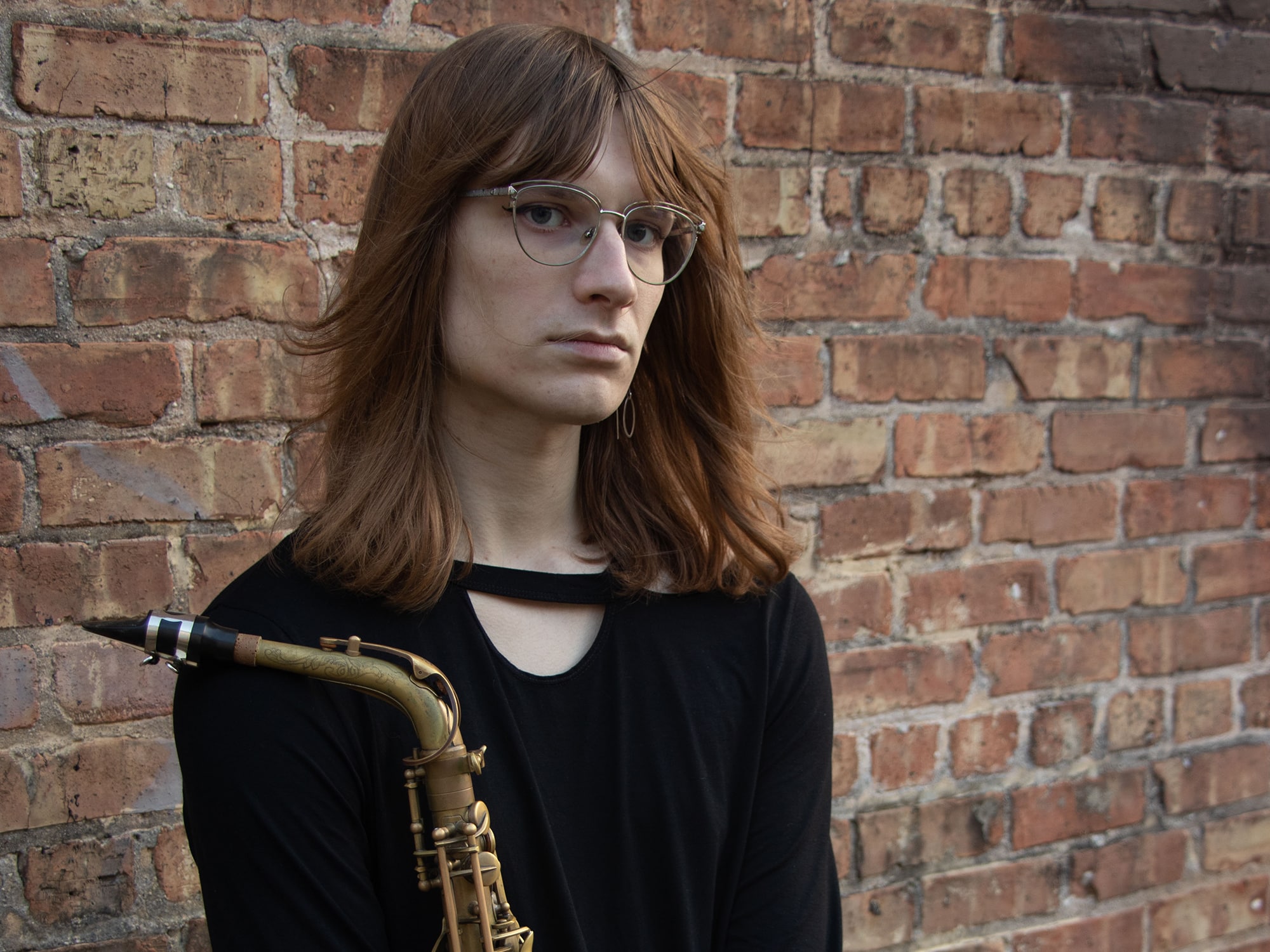 A publicity photo shows musician Maggie Cousin posing and holding a saxophone, with a brick wall in the background.
