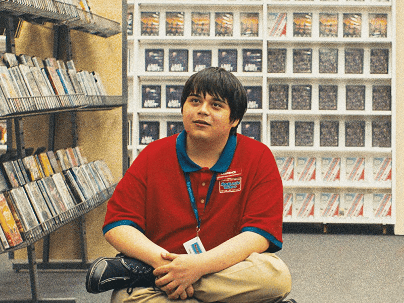 A cropped image from the "I Like Movies" poster shows actor Isaiah Lehtinen sitting on the floor in the Sequels Video rental store. He wears a red shirt with a blue collar and stares into the distance.