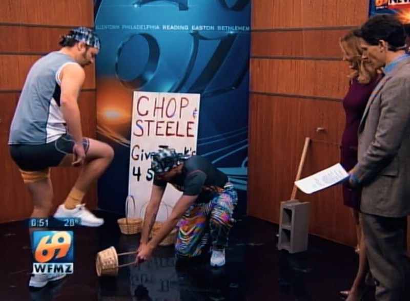 Chop (Joe Pickett) lifts his foot while Steele (Nick Prueher) holds a wicker basket on the ground to be stomped in a local news morning show studio. Two news anchors to their right watch their activity. A sign reading “Chop & Steele Give Thank’s 4 Strength’s” Pickett rests against a wall displaying the TV station behind them.