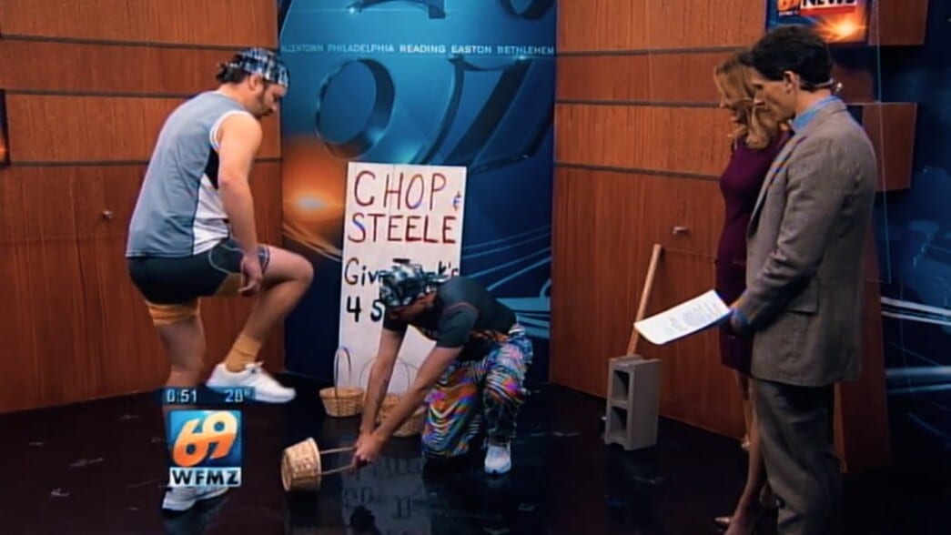 Chop (Joe Pickett) lifts his foot while Steele (Nick Prueher) holds a wicker basket on the ground to be stomped in a local news morning show studio. Two news anchors to their right watch their activity. A sign reading “Chop & Steele Give Thank’s 4 Strength’s” Pickett rests against a wall displaying the TV station behind them.