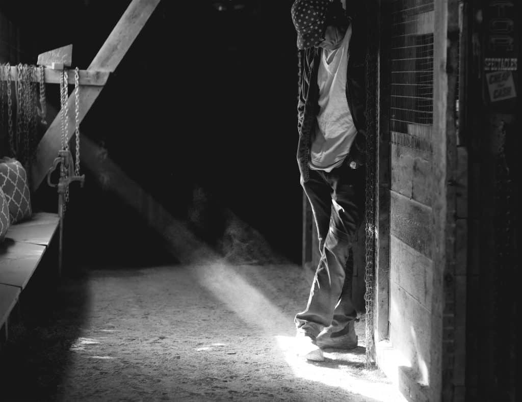 A black-and-white photograph depicts Matt Monsoor standing with his head down and legs slightly crossed while leaning against a wall in a wood shed. He's pictured to the slight left of center, wearing a jacket, work pants, a white shirt, white sneakers, and a black-and-white baseball cap that partially obscures his face. Beams of light cut in from the right and illuminate his shoes. The floor is dirt.