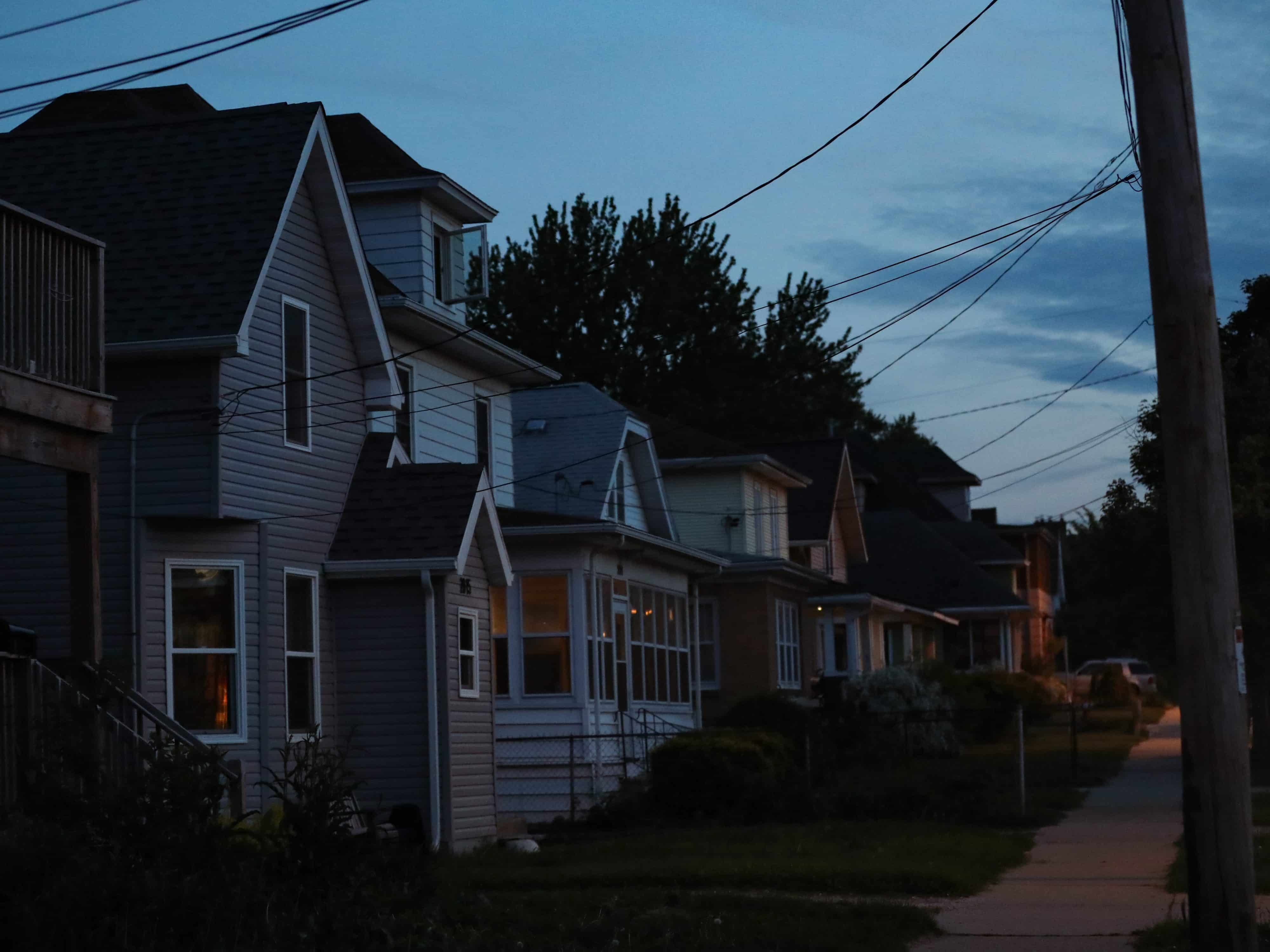 A photograph depicts a row of homes placed closely together along a sidewalk on Madison's east side. The sun is setting and there is a pale blue tint to the image.