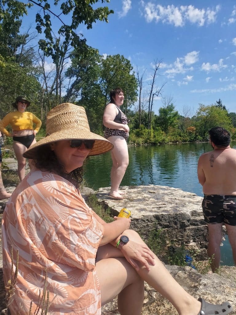 A photo of someone in a straw hat and bathing suit cover sitting by a lake. In the background, people in bathing suits laugh.