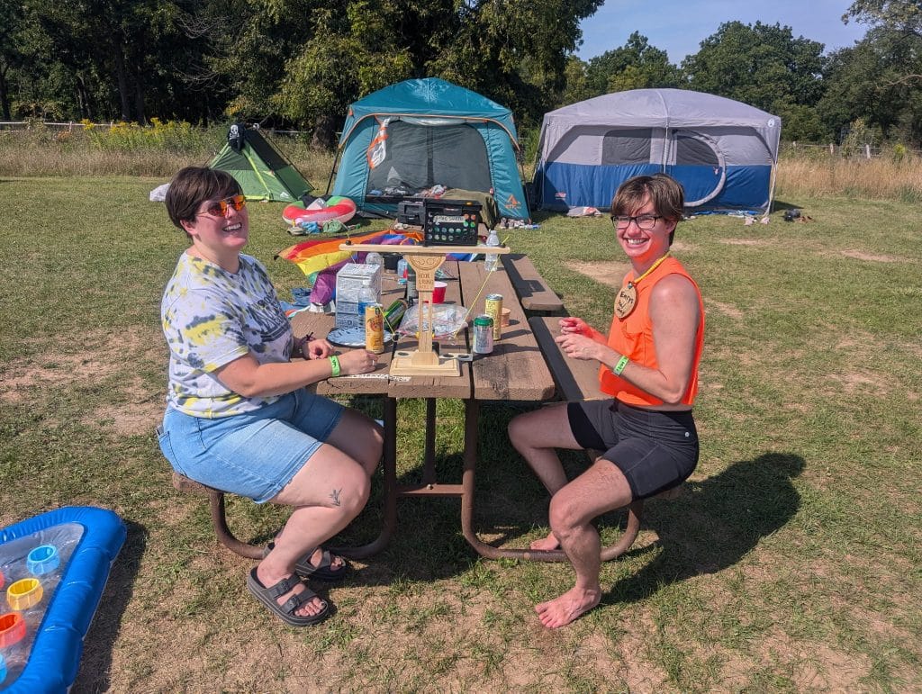 An outdoor photo of two people in summer clothes sitting opposite one another at a picnic table. They smile and face the camera. A wooden game called "Hook and Ring" sits on top of the table along with some aluminum cans and plastic bottles. There are three tents pitched in the grass in the background behind them.
