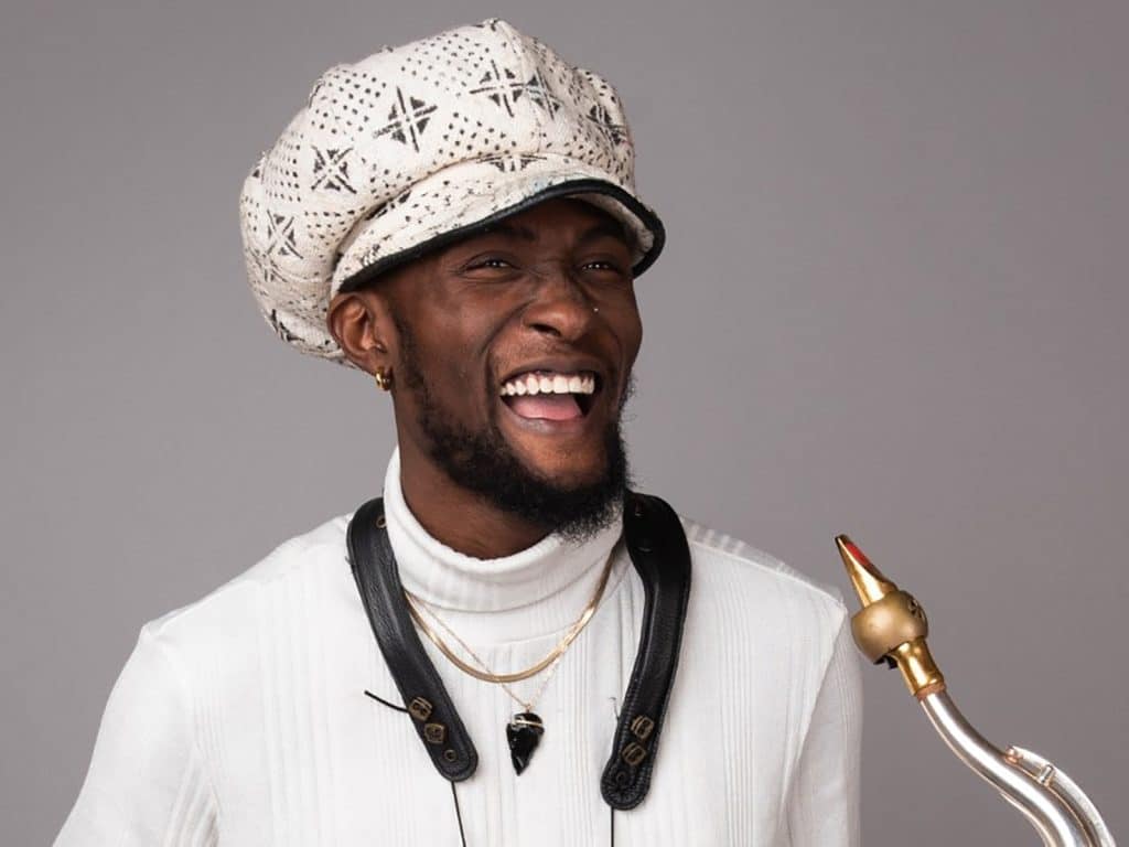 Isaiah Collier is shown in the center of the image, smiling at the camera. He's wearing a white turtleneck and an eccentric white hat. The background is gray.