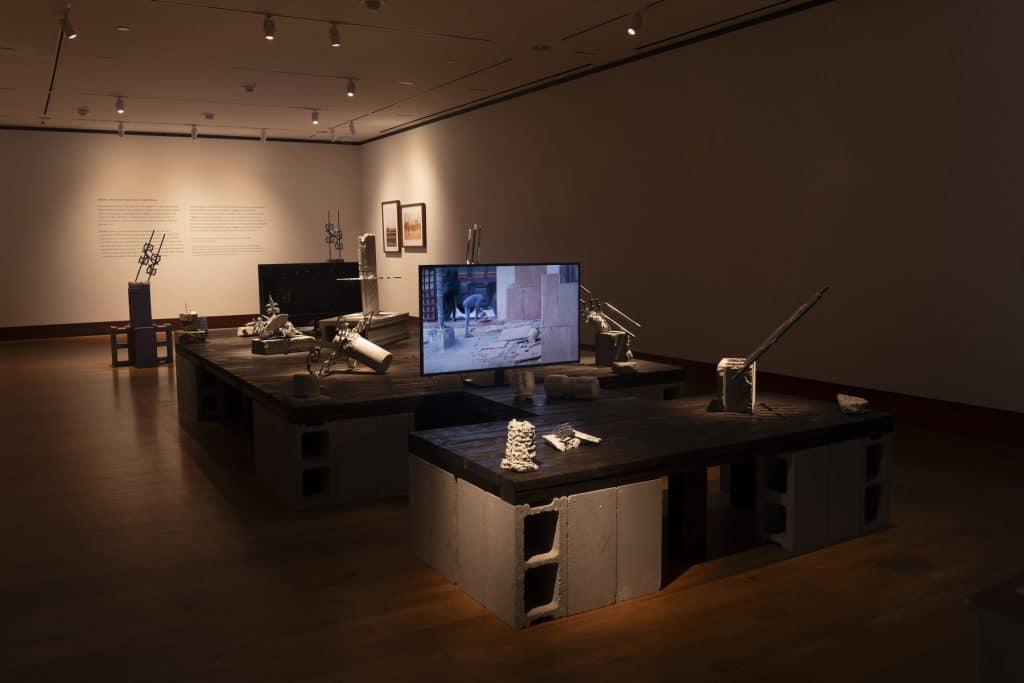 A dimly lit gallery shows various sculptures made of concrete and steel on the floor and propped up on tables made of wooden slabs and cinderblocks. A flatscreen monitor on the nearest table displays footage of a construction site. In the distance, two photos are mounted on the right-hand wall. And, to their left is Anamika Singh's artist statement about her work.