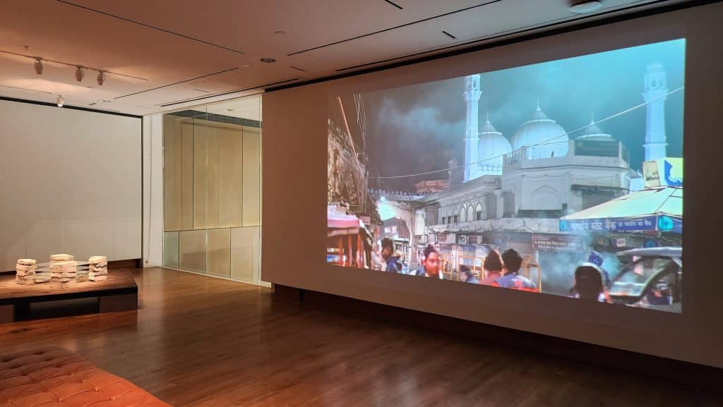 A photo displays a more spacious gallery with a white wall that is being used as a projection screen for B-roll footage taken in Faizabad and Ayodhya in the 2020s. To the immediate left of that is a clear glass window that is letting in some sunlight. In front of that, a concrete and glass sculpture sits on a small, square table low to the floor.