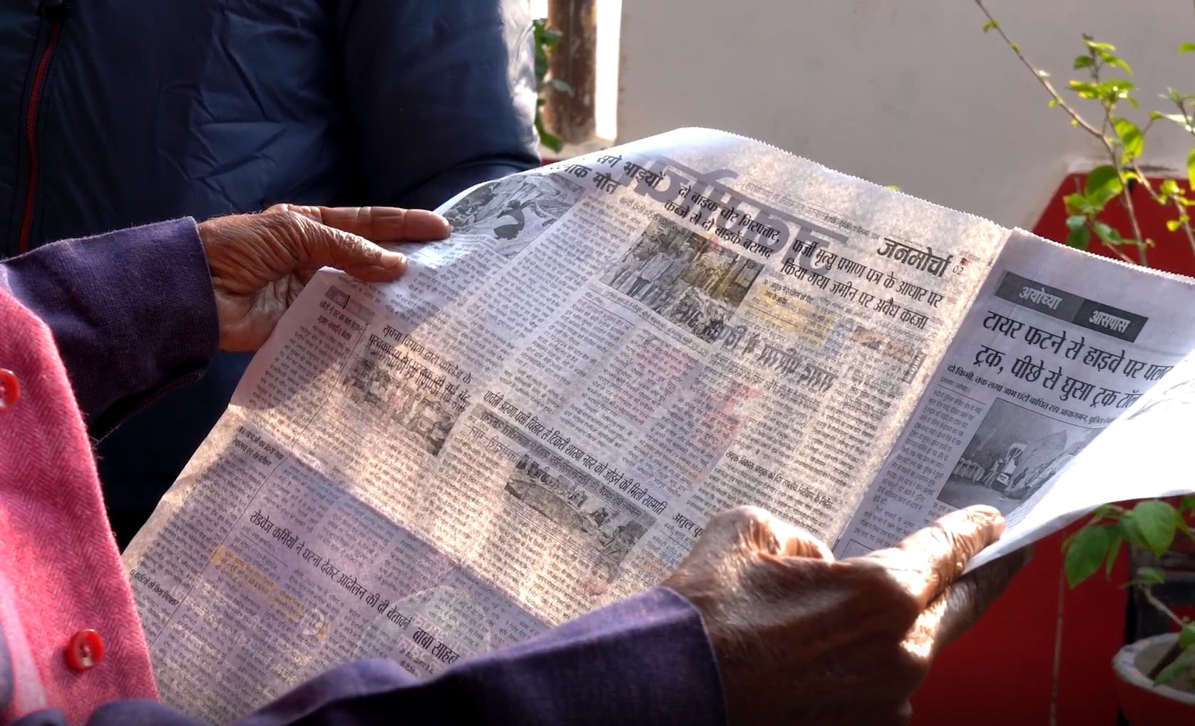 An Indian person's hands are shown at a close-up holding open a printed copy of a newspaper. Sunlight makes parts of the newsprint transparent, blurring the Hindi text on the cover page and second page that are printed on opposite sides.