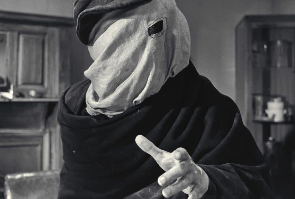 A black and white still image from the 1980 film, "The Elephant Man" shows a male figure standing in an office room. He is shrouded in dark garments and a lighter cloth that covers his entire head. A small eyehole section is cut from the lighter cloth to reveal his left eye. His left hand gestures forward.