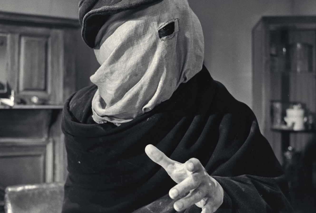 A black and white still image from the 1980 film, "The Elephant Man" shows a male figure standing in an office room. He is shrouded in dark garments and a lighter cloth that covers his entire head. A small eyehole section is cut from the lighter cloth to reveal his left eye. His left hand gestures forward.