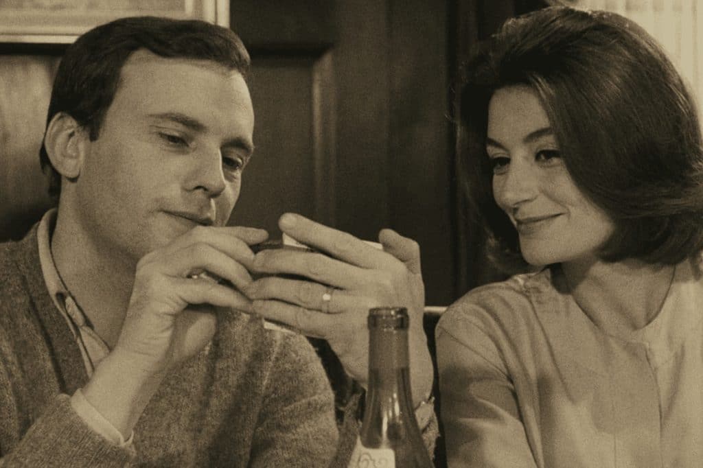 A sepia-colored still image from the 1966 French film "A Man A Woman" shows the titular man and woman sitting at a restaurant booth at a close-up. The man, on the left, holds an object up in his left hand to show the woman seated beside him. They're both wearing semi-formal clothing and smiling.