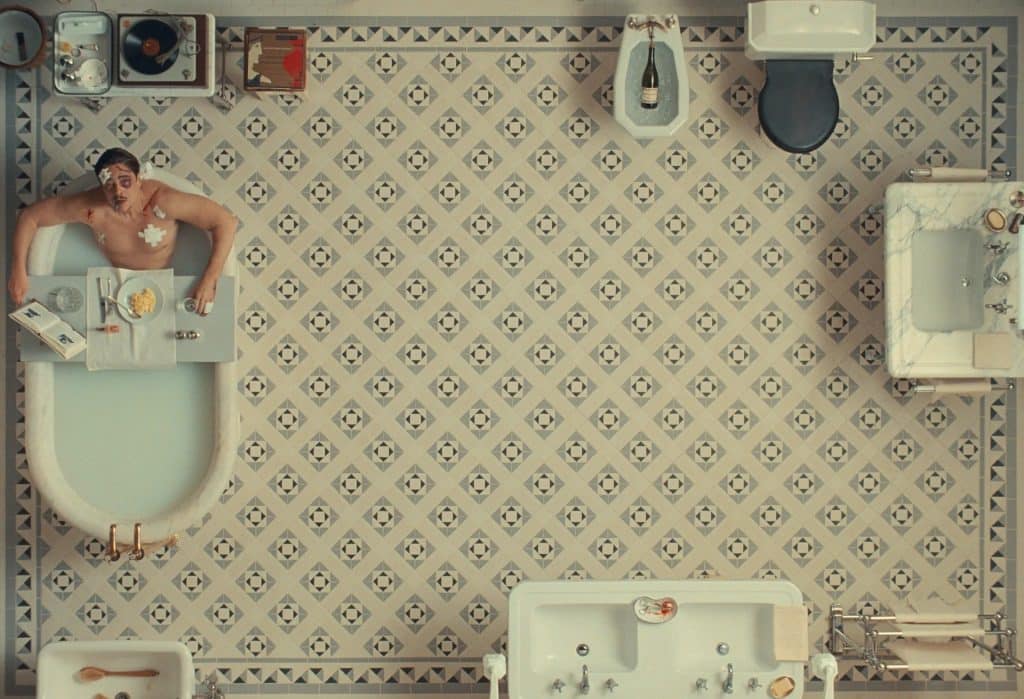 A still image from the film "The Phoenician Scheme" shows an overhead shot of a large bathroom with several sinks, a toilet, a turntable, and a bathtub all at the edges of the frame. Soaking in the bath is a man in his 50s. He has several wounds on his face and chest, and some are covered with bandages. The man stares up towards the ceiling. A caddy or tray of food is perched across the width of the bathtub.