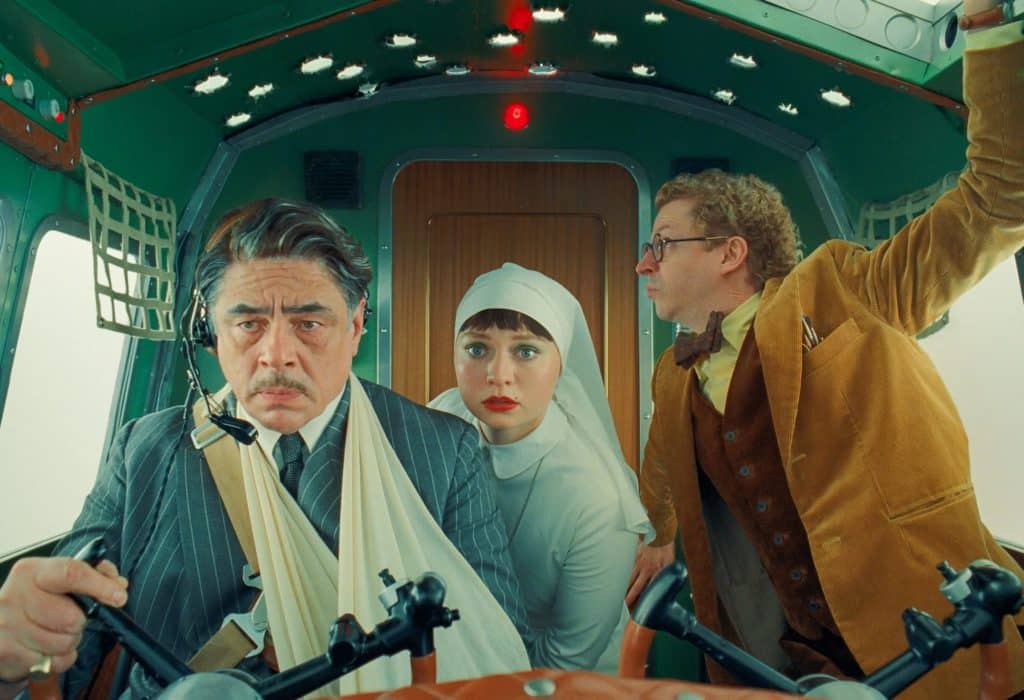 A still image from the film "The Phoenician Scheme" shows three figures aboard a small aircraft in the cramped cockpit. A man in his 50s wears a business suit, sitting at the left with a broken arm; but he's flying the plane nonetheless. To his immediate right and behind him, a younger woman crouches and stares ahead with a concerned expression. She is dressed in a white habit as a novice nun. And to her right is a slightly older man dressed in a tan corduroy suit with a vest and bowtie. He stares out the lefthand window of the plane. All their expressions reflect concern or alarm.