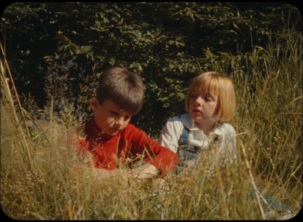 Two children sit on a hillside with tall grass on a summer's day. The boy on the right wears a red long-sleeve shirt, and the girl on the right wears a white collared shirt with blue denim overalls. The girl turns towards the boy, who has his head down as if lost in thought.