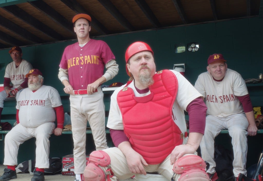 Five aging players on a recreational baseball league team named "Adler's Paint" sit in their dugout and look toward the ball field off screen. The catcher sits in the foreground near the center with protective gear on.