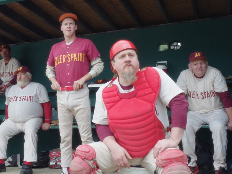 Five aging players on a recreational baseball league team named "Adler's Paint" sit in their dugout and look toward the ball field off screen. The catcher sits in the foreground near the center with protective gear on.