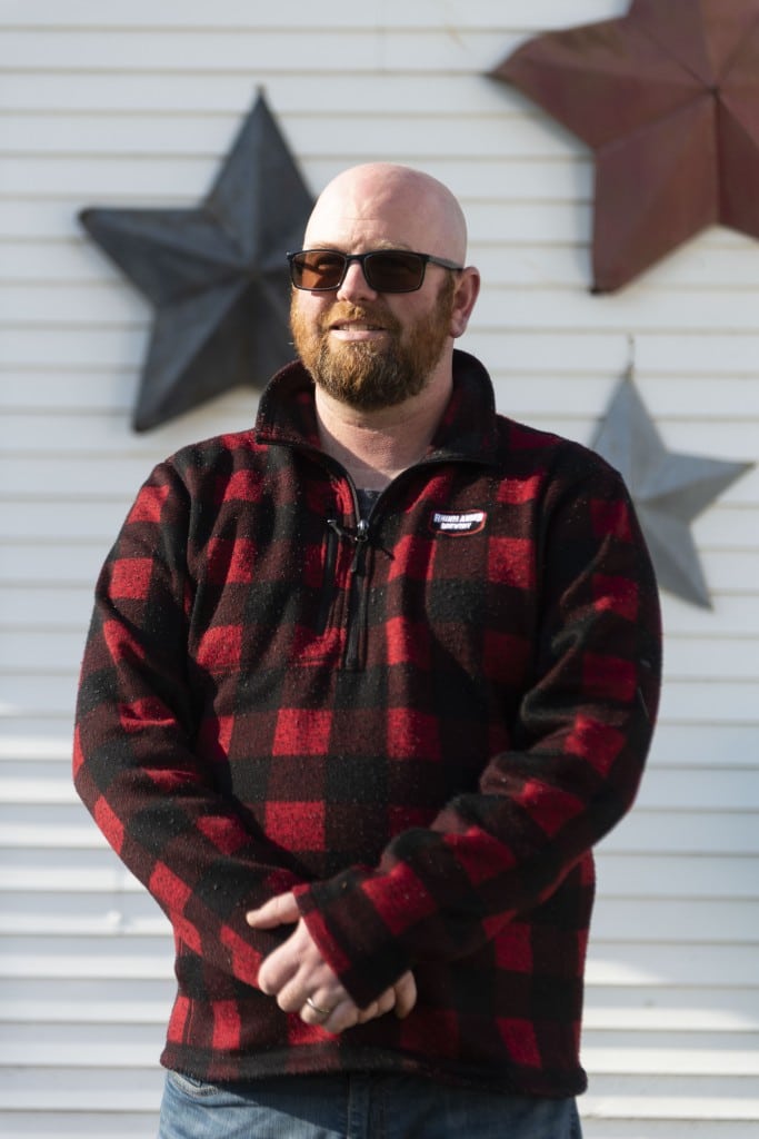 Photograph of a bearded man wearing a flannel fleece and sunglasses. He stands upright with his arms hanging down in front of him.