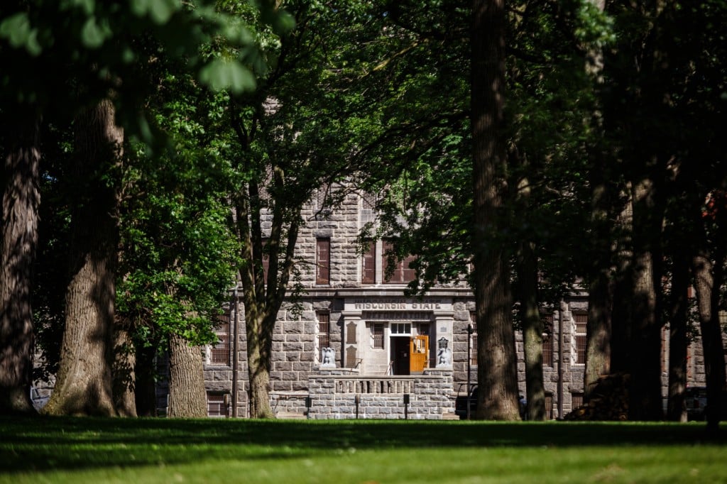 Photograph shows a large stone building in the distance with several trees in the middle ground that provide ample shade and cast shadows in the sunlight.