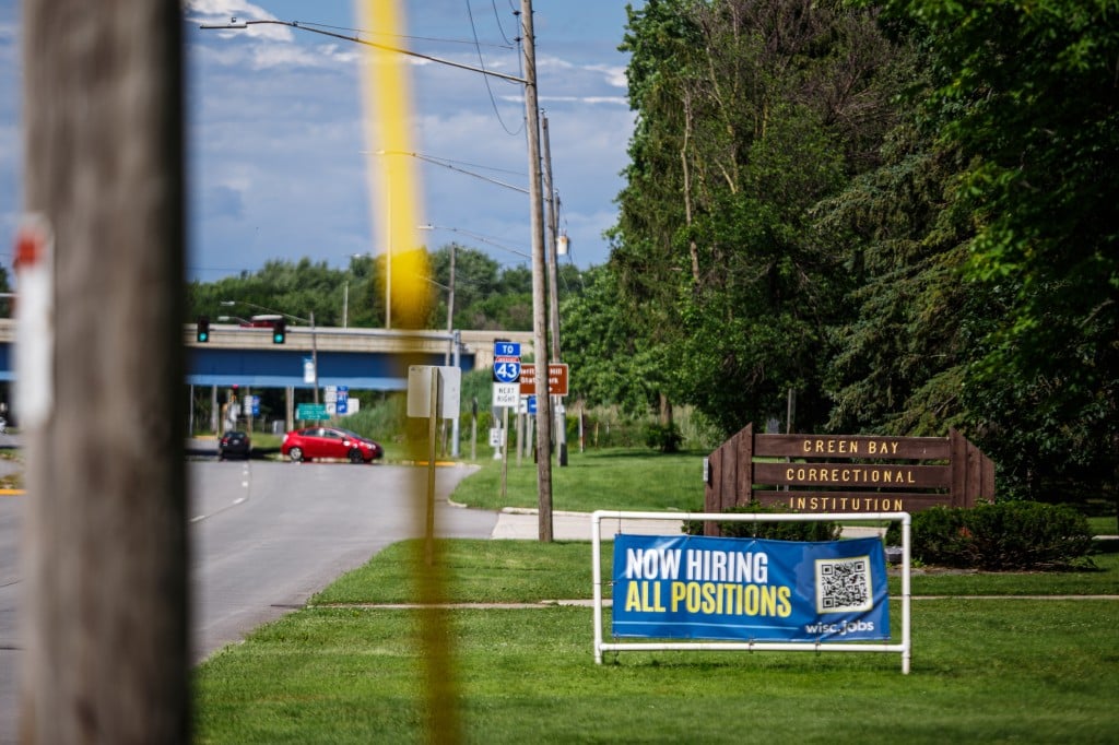 Photograph from the shoulder of a road shows a wooden sign for "Green Bay Correctional Institution" in the middle distance with a plastic sign that reads "Now Hiring All Positions" in front of it in the grass. It is a sunny, partly cloudy day.