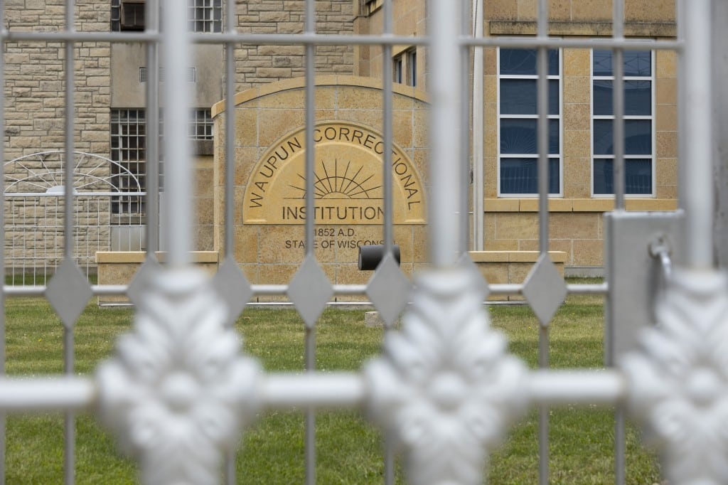 Photograph of two steel gates in front of the Waupun Correctional Institution. The tan-colored stone sign in the background sits near the building itself. The first gate at the foreground is out of focus, while the second gate and rest of the photo is in focus.