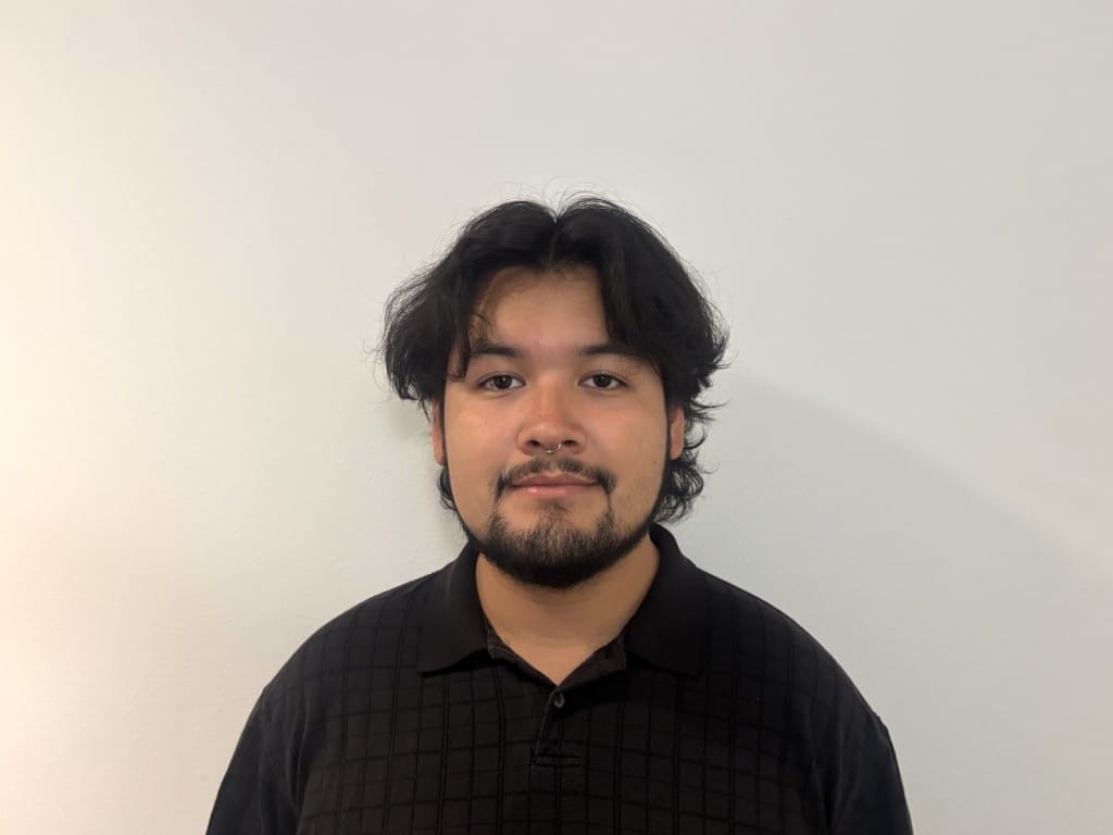 A one-quarter headshot of Tone Madison's incoming News and Politics Editor, Juan Carlos Garcia Martinez. He wears a black collared shirt, stands against an eggshell-colored wall, and faces the camera.