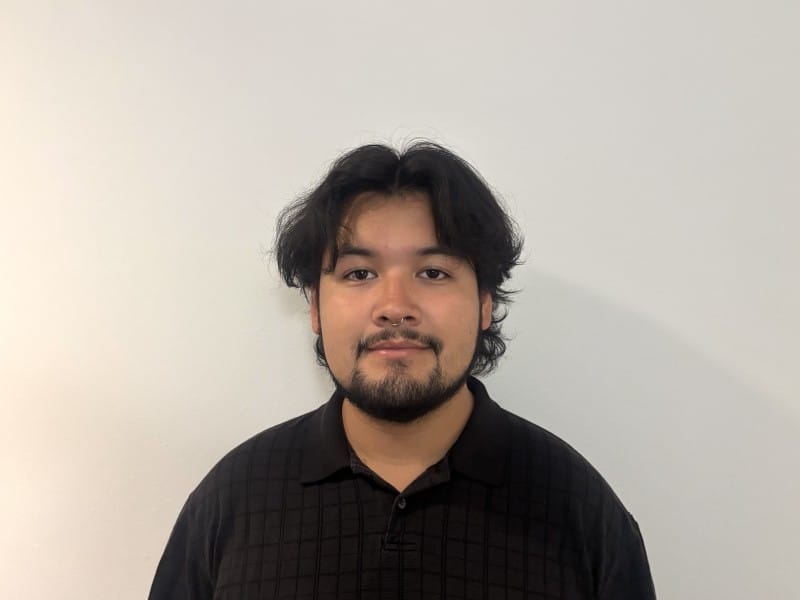 A one-quarter headshot of Tone Madison's incoming News and Politics Editor, Juan Carlos Garcia Martinez. He wears a black collared shirt, stands against an eggshell-colored wall, and faces the camera.