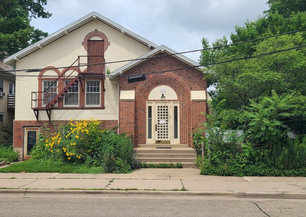 A landscape photograph of a small building with unique brick architecture. The building contains a main entrance on the ground level, up a series of five stone steps as well as an upper-level entrance to the left that has a wooden door. A steel staircase leads down from that wooden door towards a series of three windows. Various greenery and flowers are overgrowing in front of the building by the sidewalk.