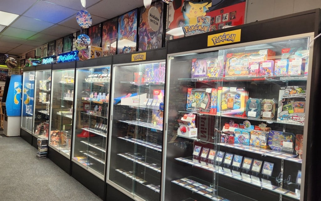 Photo shows, at a sharp angle, a series of six display cases on a store floor. The fluorescent-illuminated cases contain various collectible paraphernalia and merchandise. The first three visibly display cards and sets related to the Pokémon Trading Card Game.